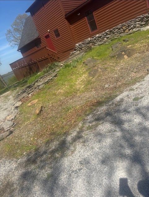 Red log cabin on hillside with gravel driveway, green grass, and wooden deck visible